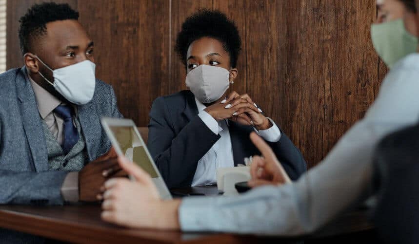 People in suits wearing face masks, sitting at a table and having a discussion.