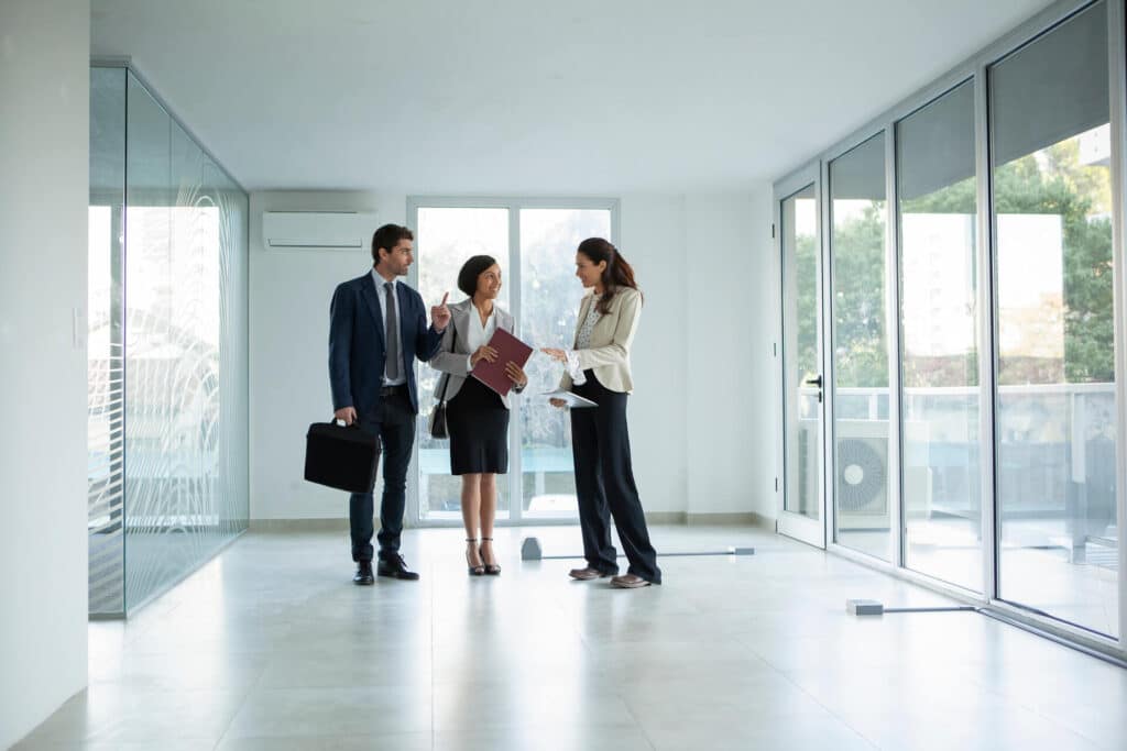 Three professionals discussing in an empty office space with large windows and natural light.