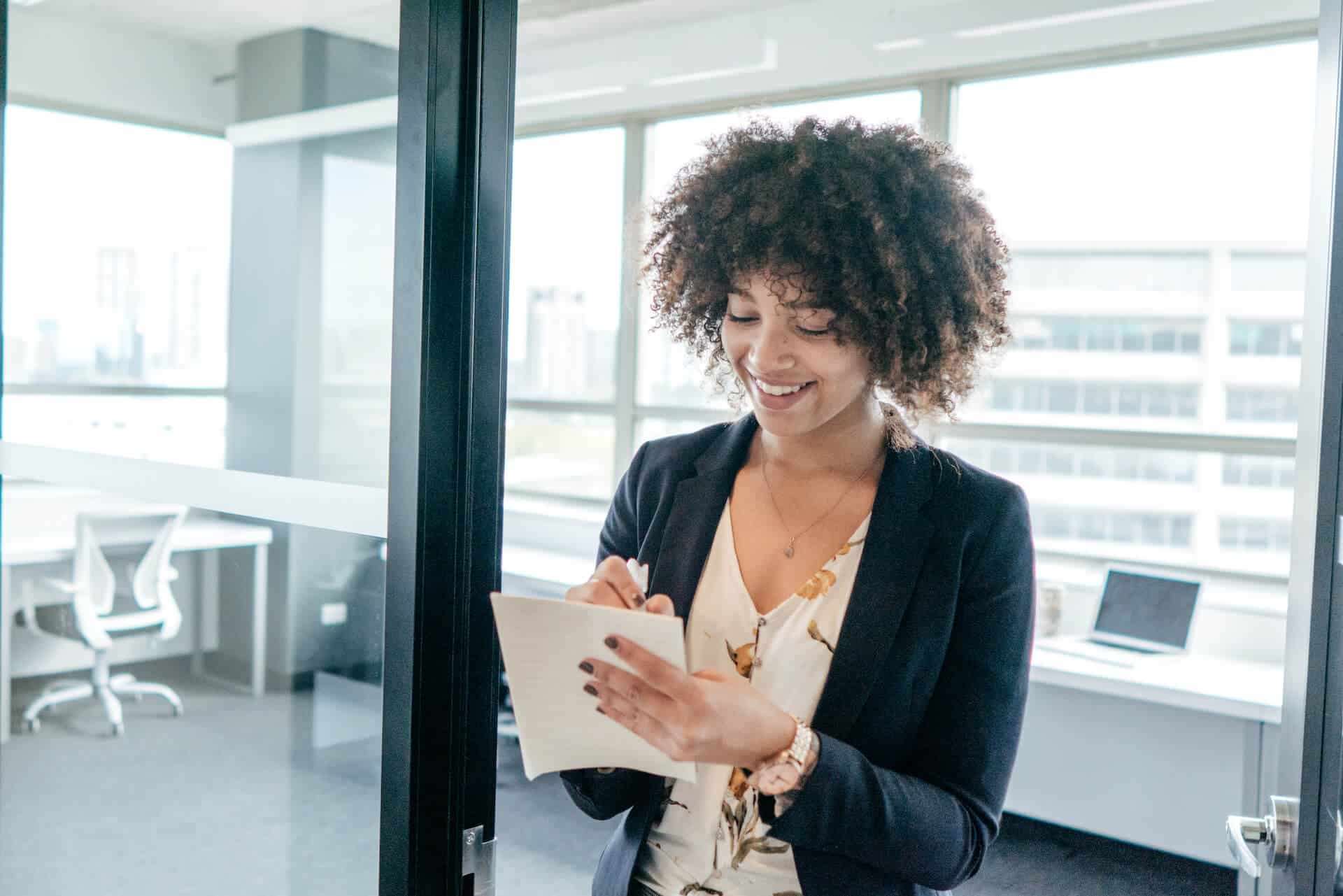 Smiling woman with curly hair writing in a notepad in a bright office.