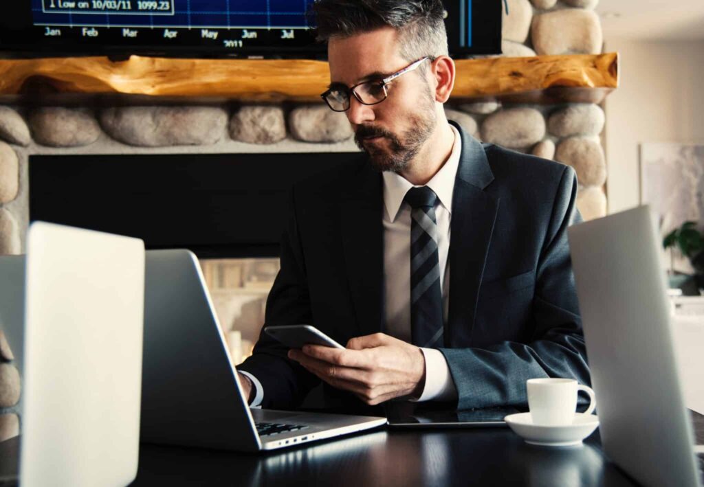 Man in a suit works on a laptop at a desk with a smartphone, coffee cup, and a stone fireplace in the background.