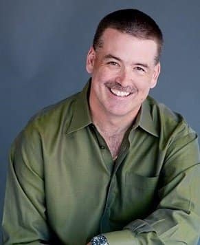 A smiling man with short hair and a mustache, wearing a green shirt, poses against a gray background.