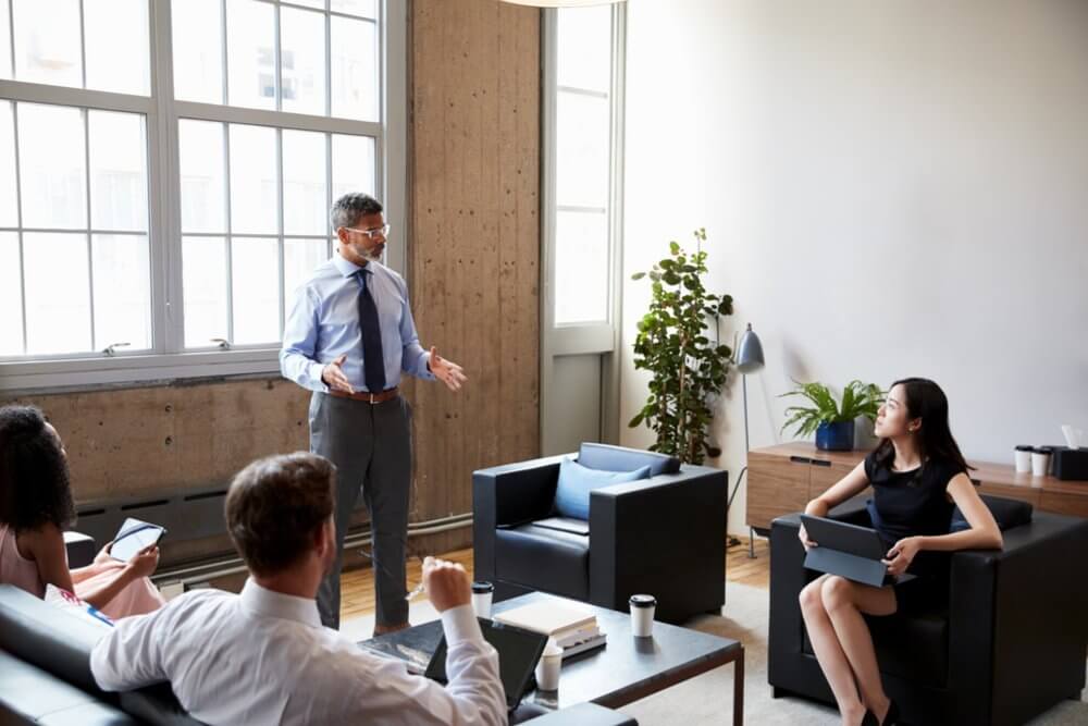 A man standing and speaking to four people seated in a modern office setting.