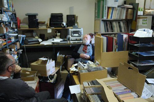 Two men in a cluttered office filled with boxes, CDs, and shelves of books.