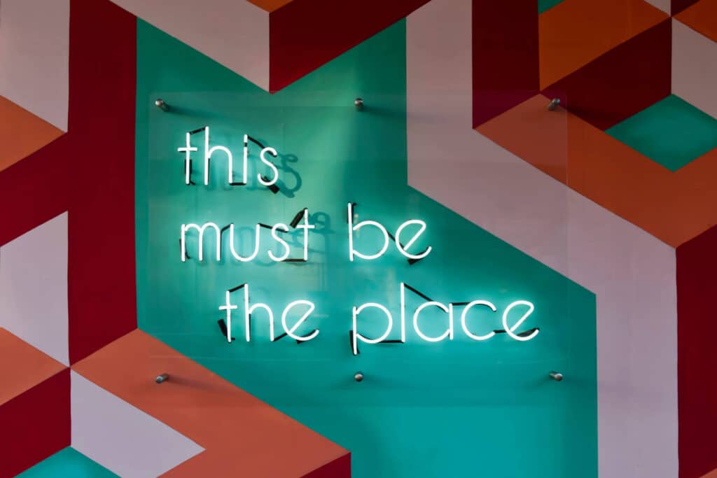 Neon sign reads "this must be the place" on a colorful geometric wall.