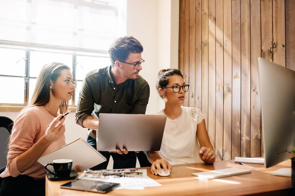 Three people working together at a desk with laptops and papers, looking at a computer monitor.