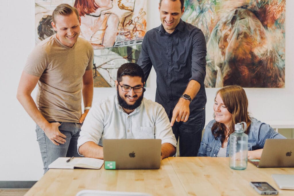 Four people happily collaborating around laptops and notebooks at a table, with artwork on the wall behind them.