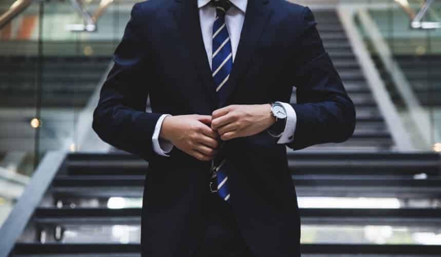 Man in a suit adjusts his jacket while standing in front of stairs indoors.