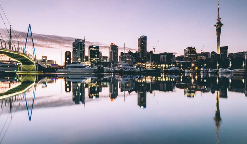 City skyline at sunset reflecting on calm water, with boats docked and a glowing bridge on the left.