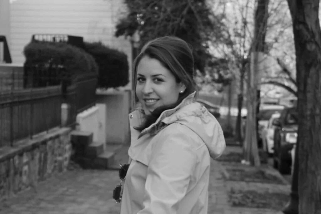 Black and white photo of a woman smiling and looking back on a leafy street.