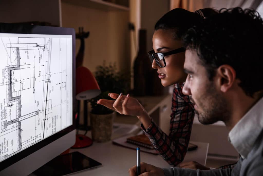 Two people discussing architectural blueprints on a computer screen in an office setting.