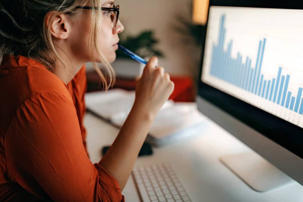 Person analyzing bar graph on computer screen at a desk, holding a pen near their chin, wearing glasses and a red shirt.