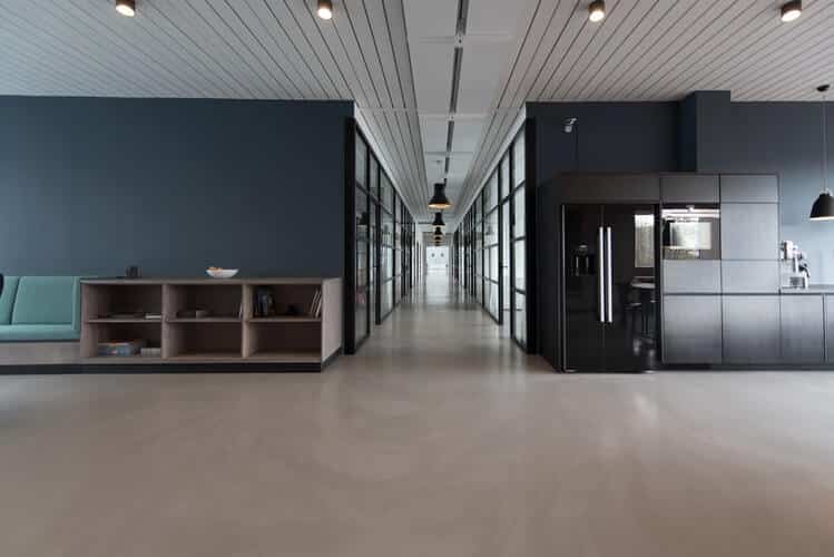 Modern office hallway with glass-walled rooms, dark cabinets, and suspended ceiling lights.
