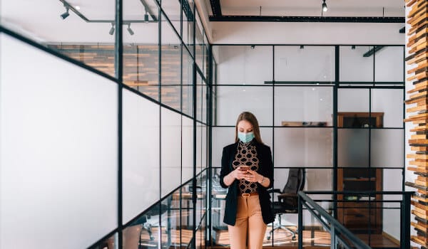 A woman wearing a mask checks her phone while walking in a modern office hallway with glass walls.