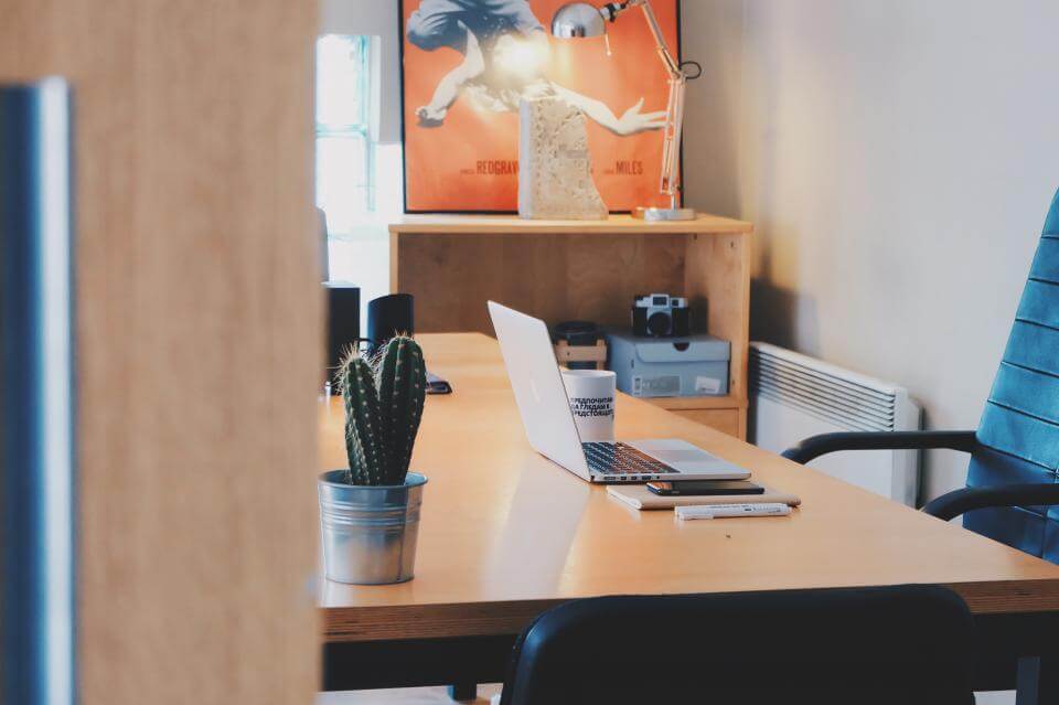 Cozy office space with a wooden desk, laptop, potted cactus, and a colorful poster on the wall.