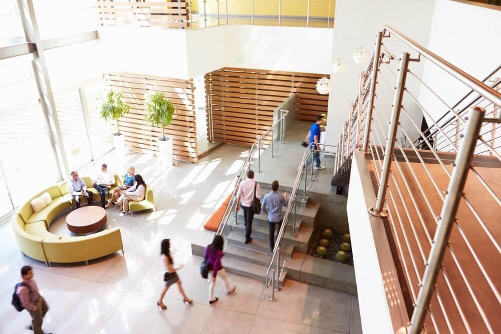 Modern office lobby with people walking and sitting, featuring a staircase, large windows, and plants.