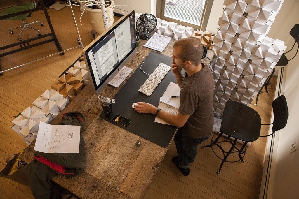 Person working at a wooden desk with a computer, surrounded by geometric paper models in a bright room.