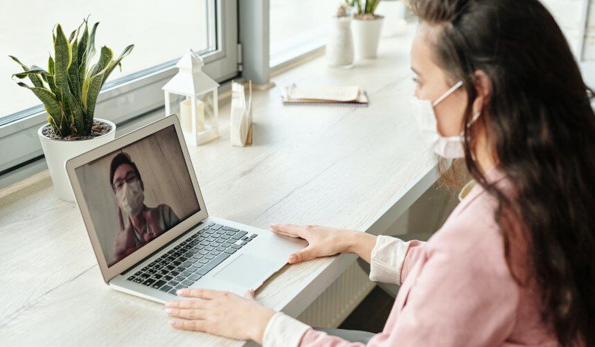 Woman in mask video calling a person on a laptop near a window with potted plants.