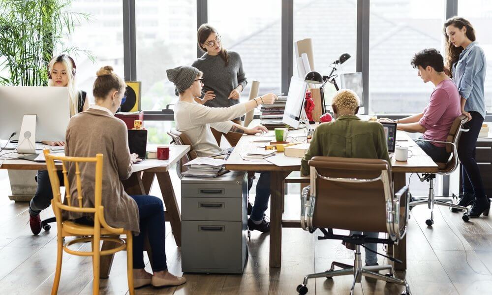 A group of people collaborating at a bright, modern office with laptops, desks, and large windows.