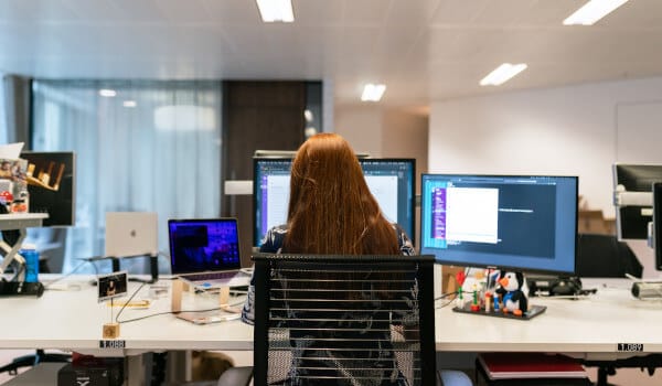 Person with long hair working at a desk with multiple computer screens in a modern office setting.