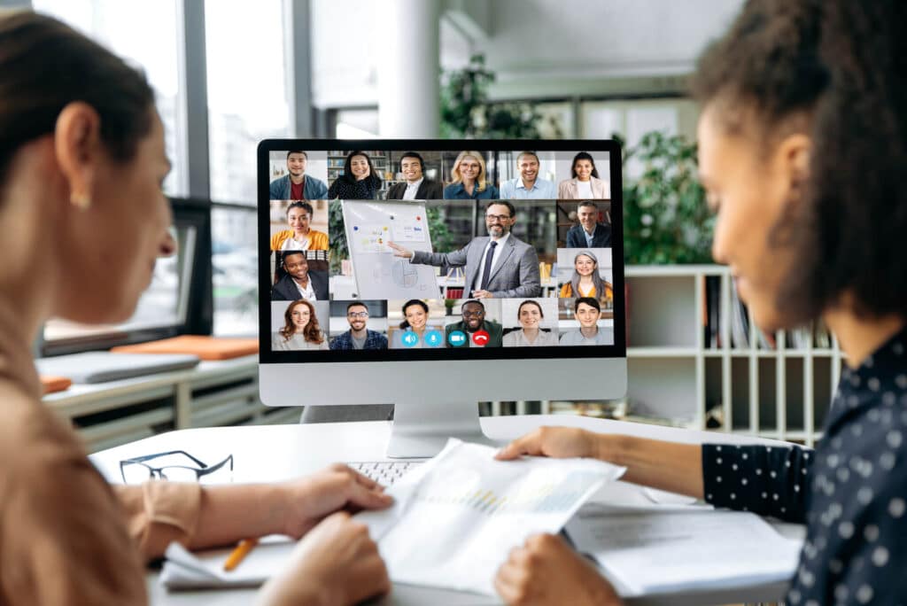Two people in a video conference, looking at documents. A virtual meeting with many participants is on the computer screen.