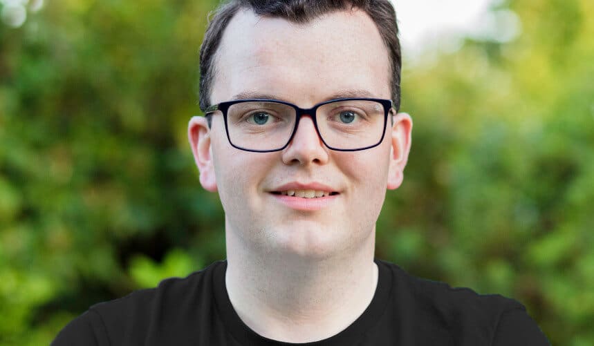 A person with glasses smiles outdoors, with green foliage in the background.
