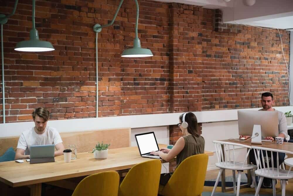 Three people working on laptops at a shared table in a modern office with brick walls and pendant lights.