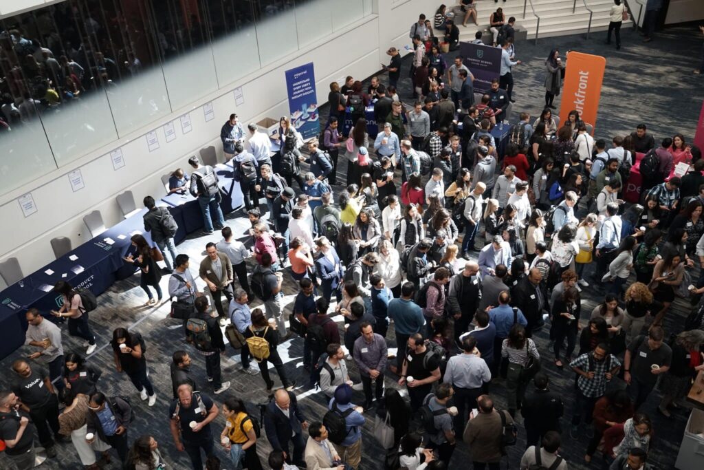 A large crowd at a conference, mingling near booths and tables in a bright, spacious hall.