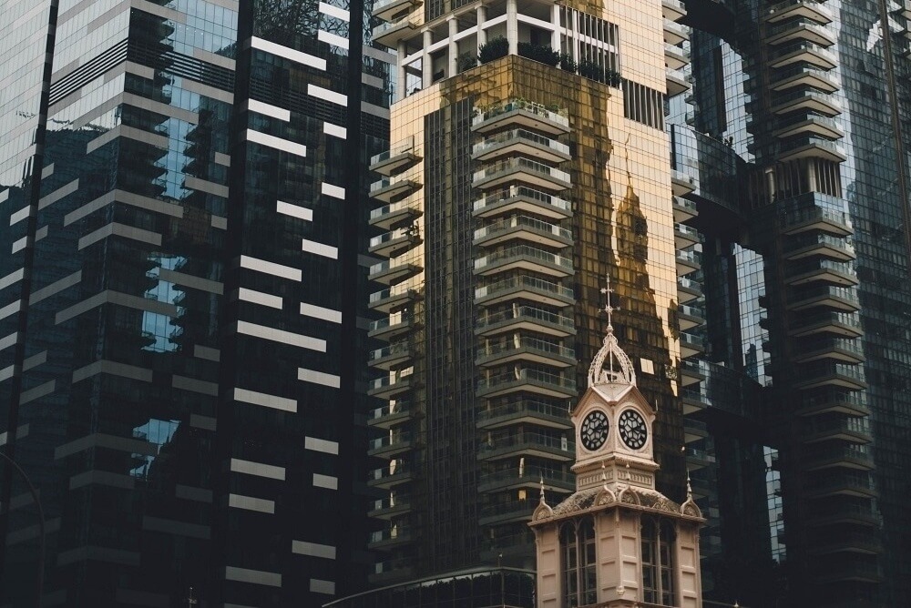 Clock tower against modern glass skyscrapers with reflective windows and geometric patterns.