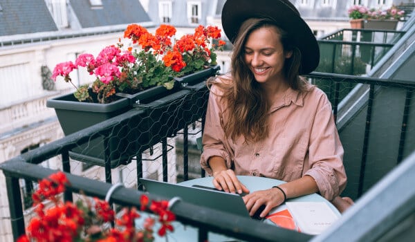 Woman in hat smiles while using a laptop on a balcony with flowers in planter boxes.