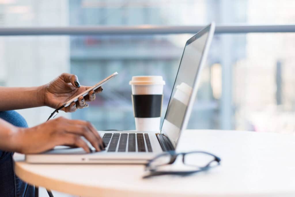 Person using a laptop and holding a smartphone at a table with a coffee cup and glasses.