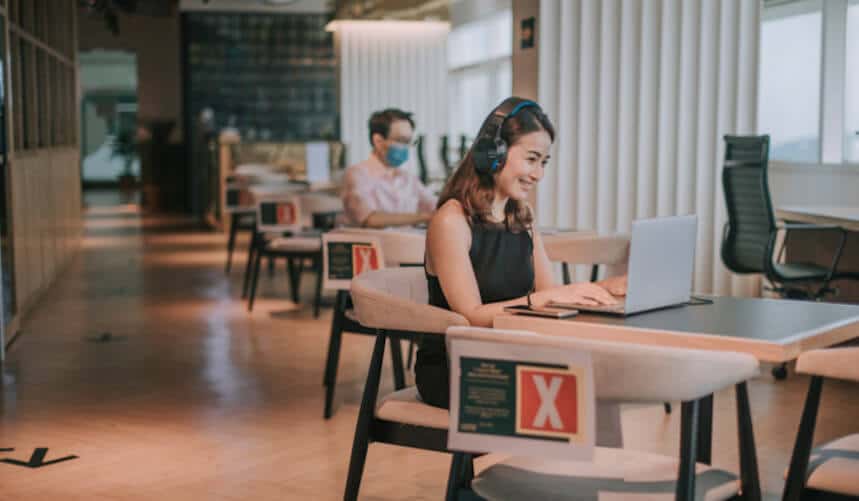 Woman in headphones working on a laptop in an office, with socially distanced seating and safety signs.