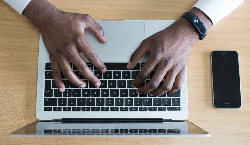 Person typing on a laptop with a smartphone and fitness tracker visible on a wooden table.