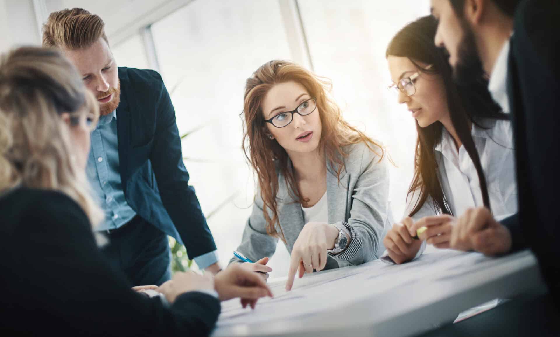 Five colleagues discussing documents around a table in a bright office setting.