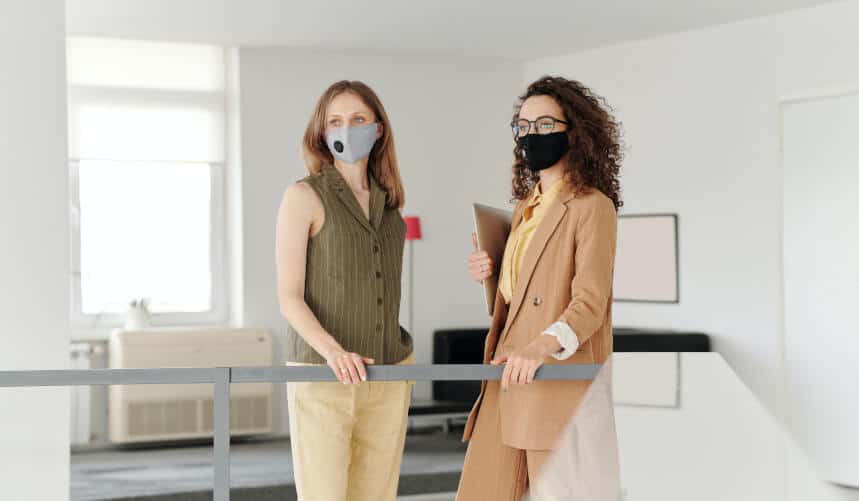 Two women wearing masks stand indoors next to a railing, one holding a laptop.