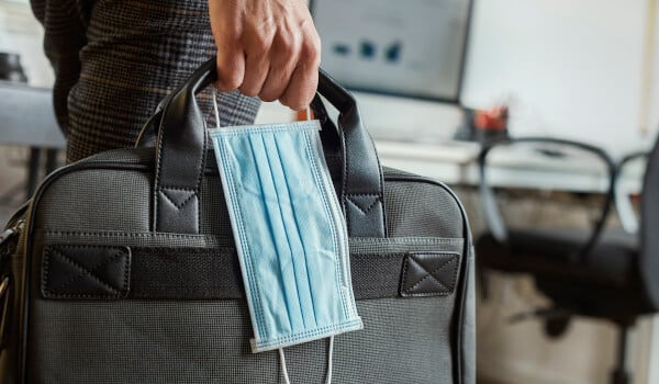 A hand holds a black briefcase with a blue surgical mask hanging in an office setting.