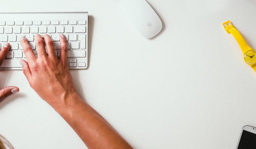 Hands typing on a white keyboard with a mouse nearby, and a yellow watch on a white table.