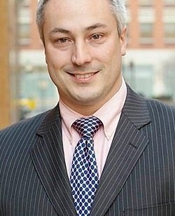 Smiling man in a pinstripe suit and patterned tie stands outdoors with a blurred city background.