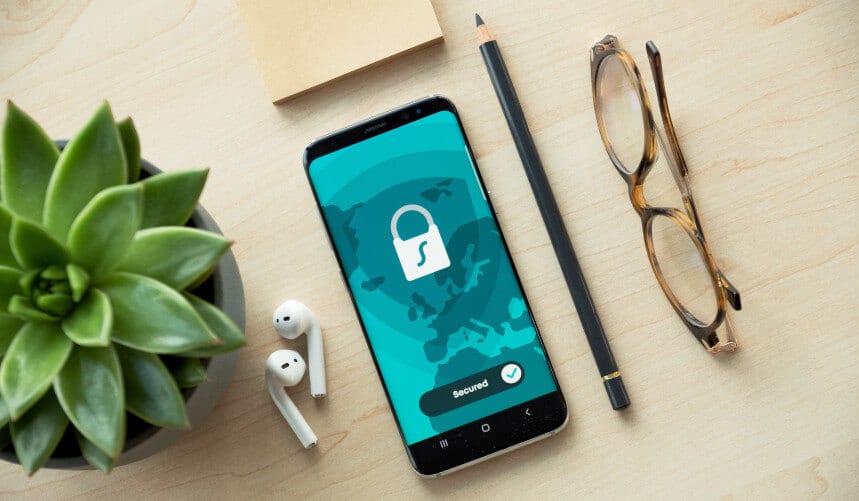 Smartphone with a secured screen beside a plant, earphones, pencil, glasses, and a notepad on a wooden surface.