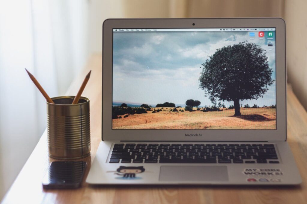Laptop on a wooden desk with a landscape wallpaper. A pen holder and a smartphone are beside it.
