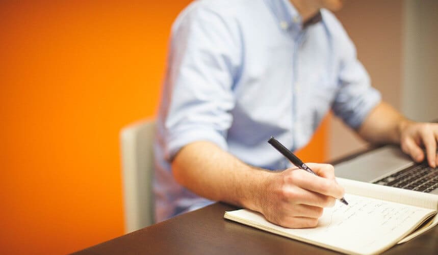Person writing in notebook and using a laptop at a desk with an orange background.