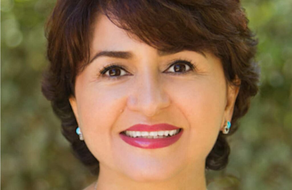 Woman smiling in front of a blurred outdoor background. She has short dark hair and wears bright lipstick.