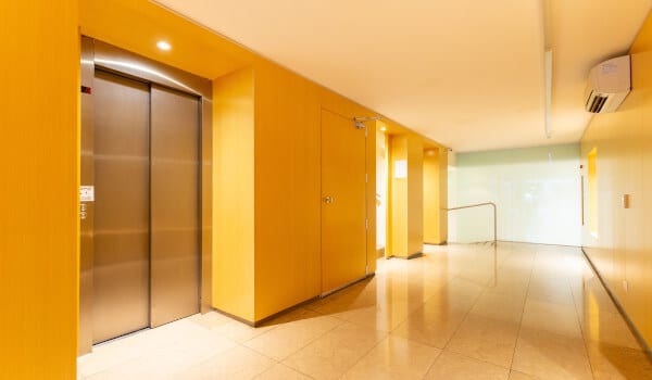 Yellow-walled corridor with elevators, beige tile flooring, and a metal handrail on the right.