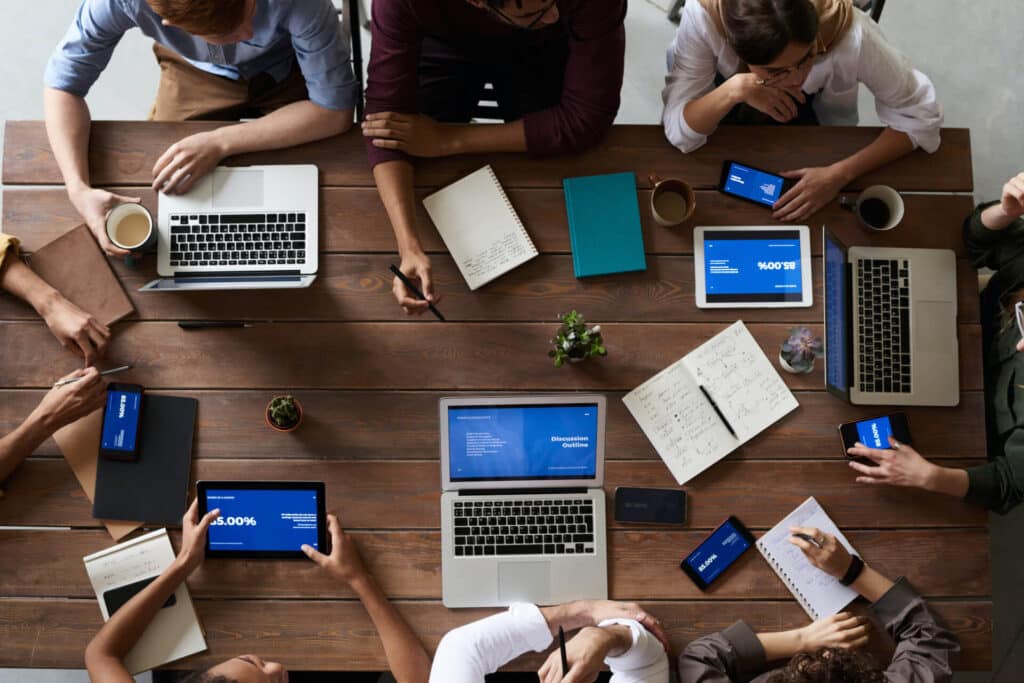 People sitting around a wooden table with laptops, tablets, notebooks, and coffee, engaged in a meeting or discussion.