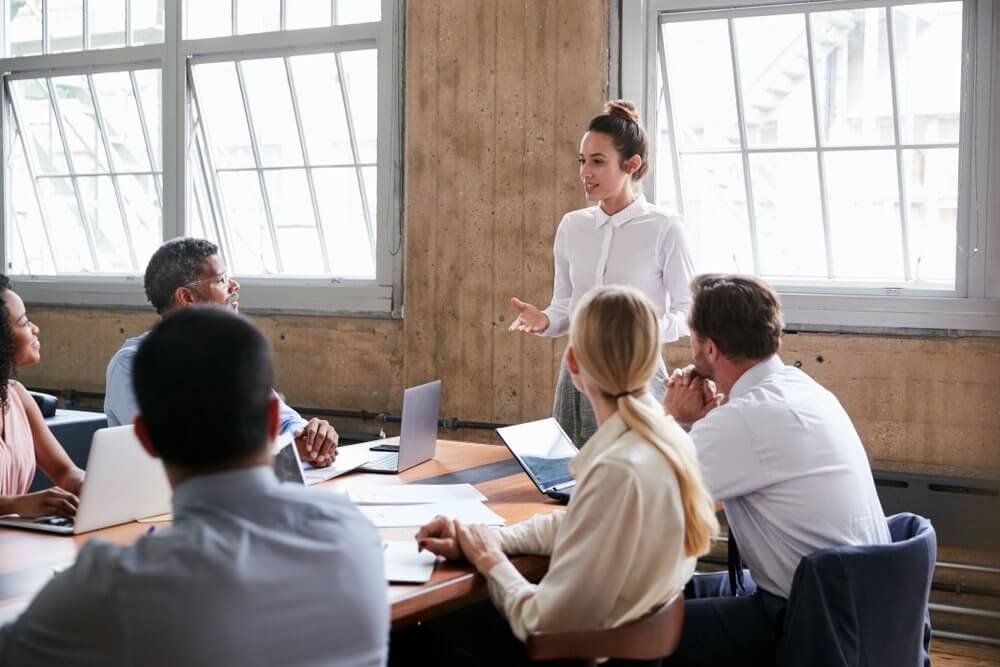 Woman standing and speaking to a group of seated colleagues in a conference room.