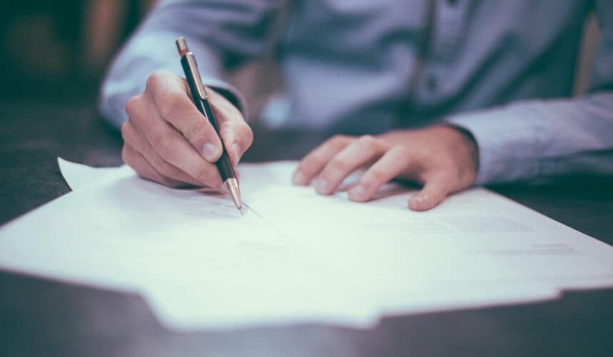 Person in a blue shirt writing on papers with a pen at a desk.