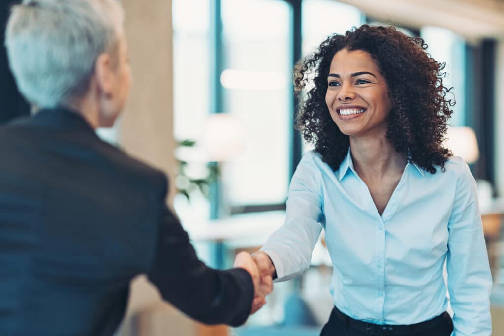 Two women shaking hands in a bright office, one smiling in a light blue shirt, the other with short gray hair.