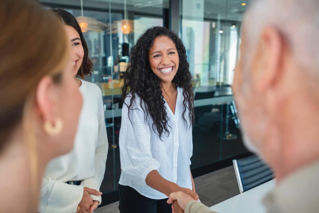 A woman smiles while shaking hands with a person in an office; two others stand nearby, also smiling.