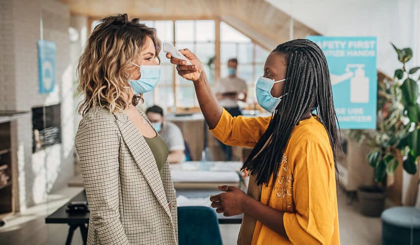 Two women in masks, one checking the other's temperature with a thermometer in an office setting.
