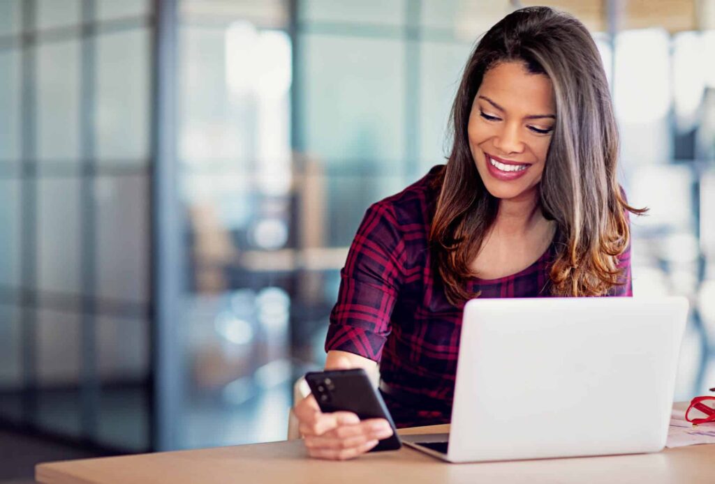 Woman looking at her phone on the side while sitting behind her laptop screen.