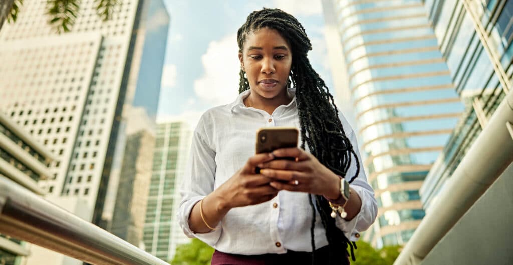 Woman with long braids using a smartphone outside, with tall modern buildings in the background.